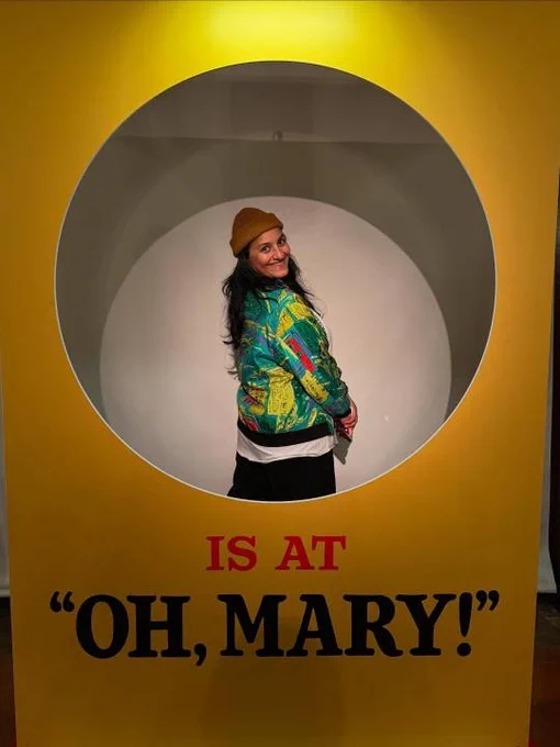 Madeleine Sami stands within a yellow circle that says 'Is At "Oh, Mary!"'