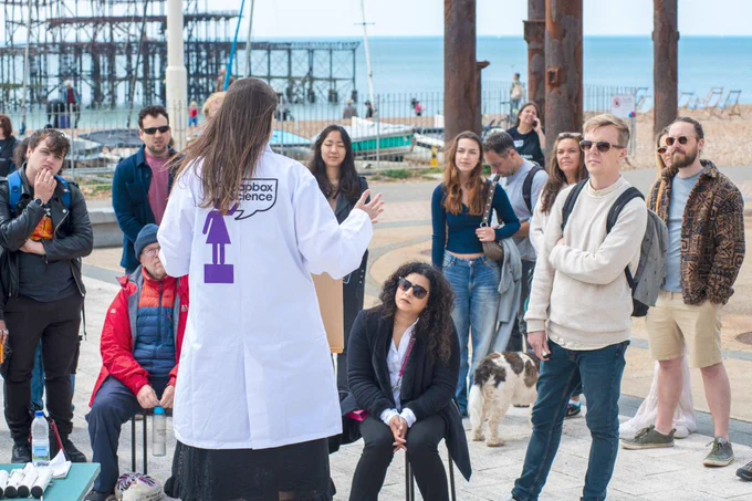 Lady wearing a white lab coat talking to a group of visitors along the seafront in front of Brighton old pier