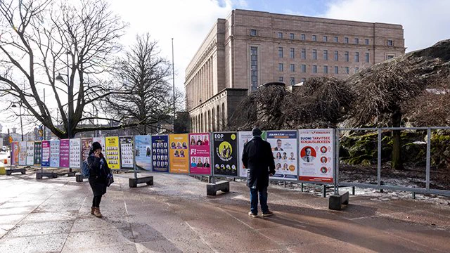 Kuvituskuvassa vaalien ulkomainoksia telineessä, joita katselemassa kaksi henkilöä. Taustalla näkyy eduskuntatalo.