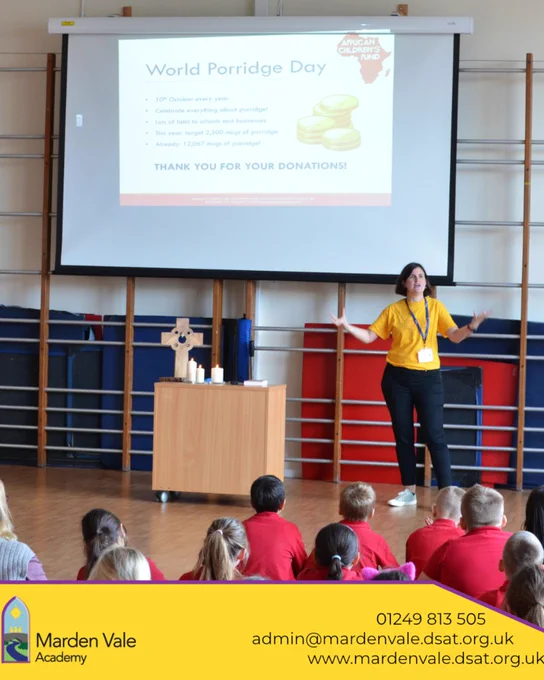 ​A lady in a yellow t-shirt standing at the front of a school hall, beneath a big screen which has a presentation on it. The backs of children in red school uniforms are looking towards the lady and the screen.