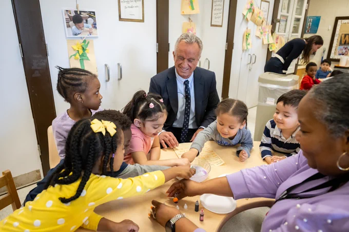 Group of children sitting around a table with Secretary Kennedy, engaging in a craft activity in a classroom setting.