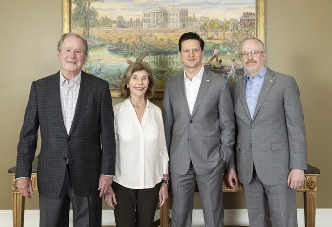 Four adults stand in front of a painting of a large building, likely a government or historic structure. From left to right, former President George W. Bush, Laura Bush, Shilo Brooks and a man in a suit with a lapel pin are pictured.