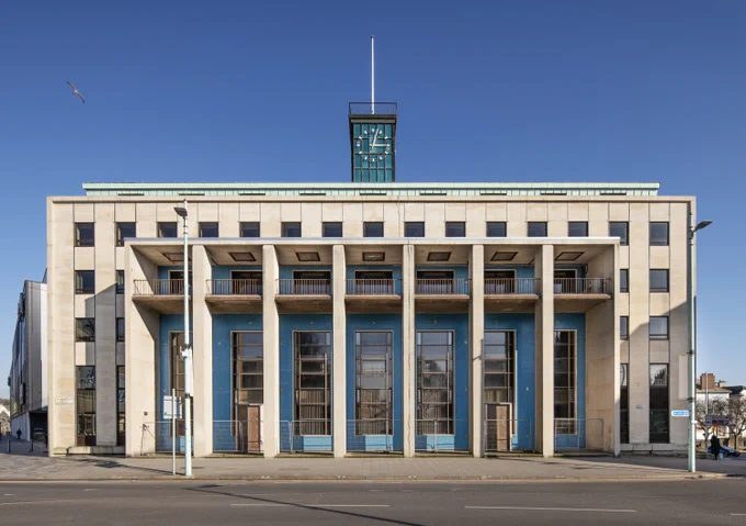 A photograph of a modernist building with tall columns, blue doors, and a clock tower showing 12:15 under a clear blue sky.