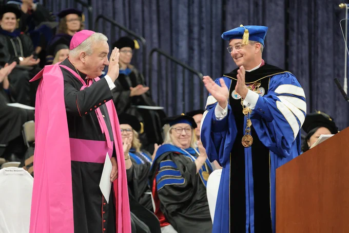 Omaha Archbishop the Most Reverend Michael McGovern waves to the crowd at Creighton University's commencement ceremonies. Creighton President the Rev. Daniel S. Hendrickson, SJ, PhD, is nearby clapping.