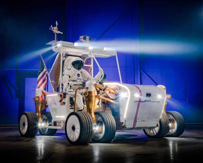 The Lunar Terrain Vehicle Ground Test Unit gleams inside of an enclosed space. An astronaut figure sits in the driver’s seat. An American flag is strapped to the back of the vehicle. Credit: NASA/Bill Stafford and Helen Arase Vargas