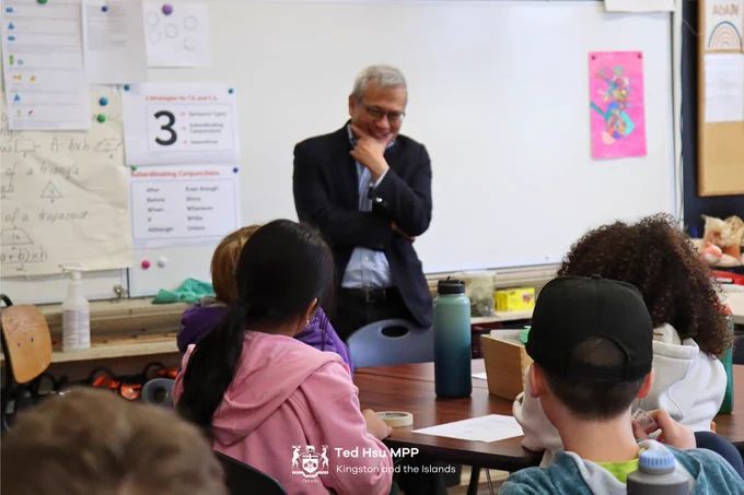 MPP Hsu standing in front of a classroom of children, he is standing smiling with his hand on his chin in a 'thinking' position.
