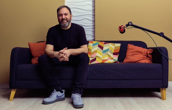 Simon Jones is sitting on a dark blue sofa with orange cushions and a podcast mic to his left.