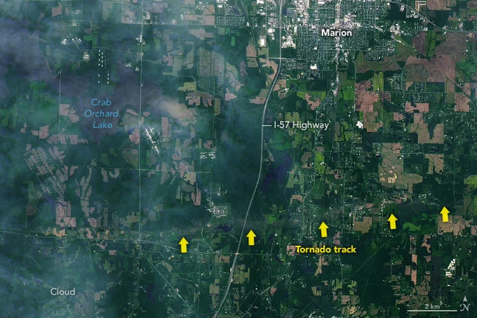 A satellite image shows a patchwork of green spaces in southern Illinois. A tornado track cuts through the vegetation from west to east. Large yellow arrows have been added to point to the tornado track. Marion, Illinois is visible at the top of the image and the I-57 Highway cuts through north to south.