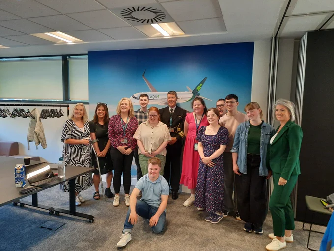 A group of TCPID students and staff with colleagues from Aer Lingus standing in a room in front of an image of an Aer Lingus plane. An Aer Lingus pilot is in the middle of the group.  