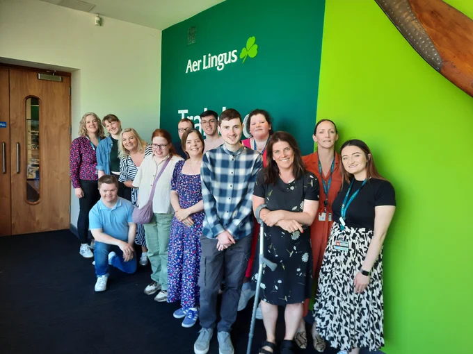 A group of TCPID students and staff standing with colleagues from Aer Lingus in front of a large green wall with the Aer Lingus logo on it.  