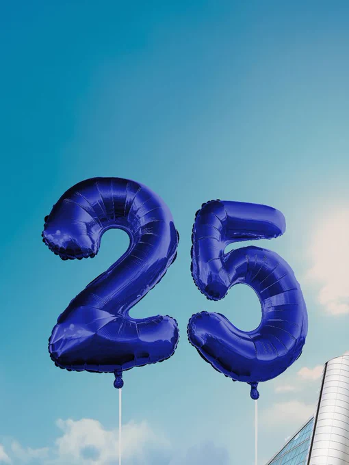 Two large blue numeral balloons, "2" and "5," floating against a clear blue sky with a small section of the Xscape Milton Keynes building visible in the background.
