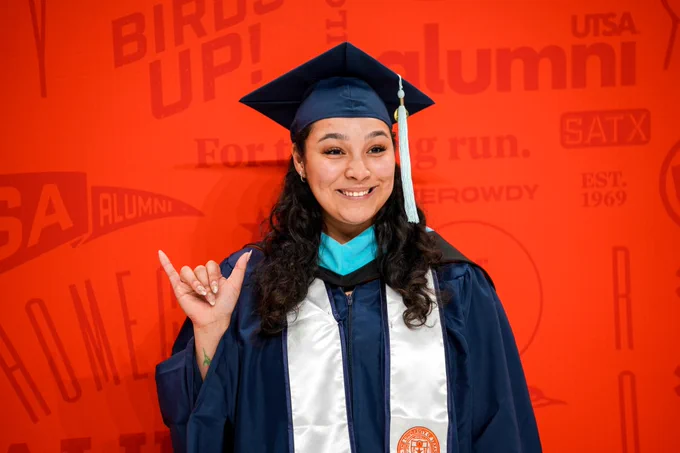 Graduate in cap and gown giving the 'birds up' hand sign in front of a University of Texas at San Antonio alumni banner.
