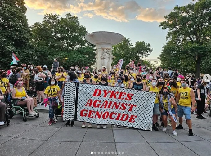 A group of figures standing, kneeling and sitting in wheelchairs outdoors, wearing masks, holding a sign reading "Dykes Against Genocide." They're standing on a gray sidewalk and there are trees in the background.