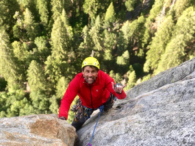Erik climbing El Capitan wearing a bright red jacket with climbing gear — ropes and carabiners— attached at the hip