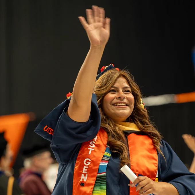 Person in graduation attire waving, with a stole marked "UTSA" and a serape sash.