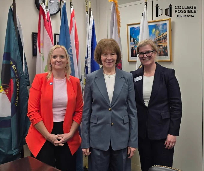 Three individuals posing for a photo in an office with various flags and a sign in the background.