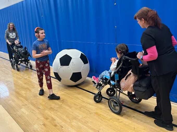 a woman pushing a child in a chair on the left side of the picture moves toward a large soccer ball. a child with headphones stands next to the ball, and on the right side another woman pushing a child in a chair moves toward it also. they are on a gym floor, behind them is a blue wall.