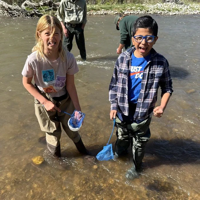 a girl and a boy in waders and boots stand in a shallow river with nets. the girl makes a disgusted face, the boy looks excited. there are two adults behind them in the river.