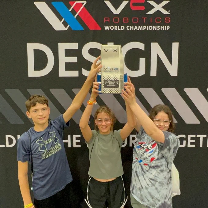 three students hold up a trophy in front of a wall with a logo and wording. 