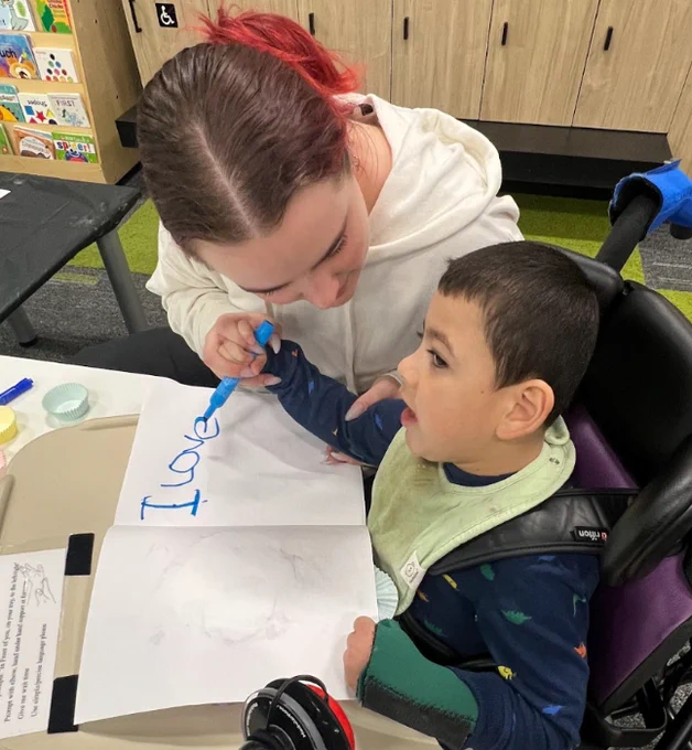 a woman helps a child in a chair write on a paper with a blue marker. the paper already says "i love"