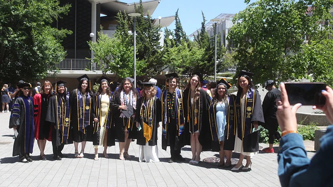 nine undergraduates lined up next to each other with two faculty members on the far right all smiling at a person taking a picture with a phone