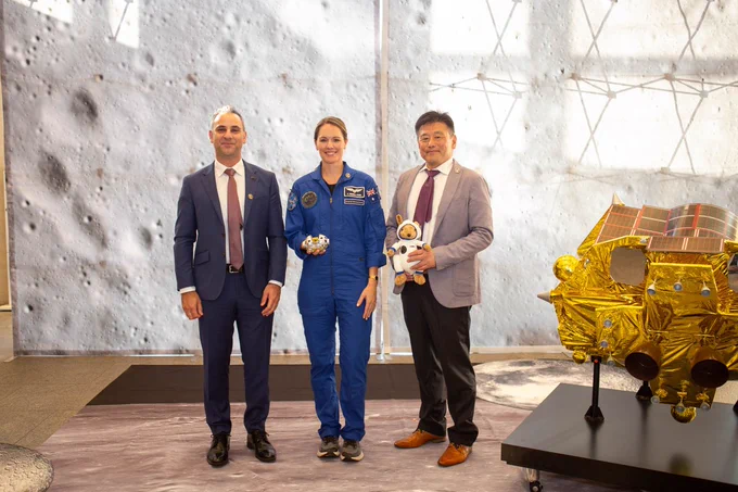 ASA Head Enrico Palermo, Astronaut Katherine Bennell-Pegg and ISAS DG Fujimoto Masaki stand in front of a large poster of the Moon's surface, and next to a model of the SLIM lander. Ms Bennell-Pegg is holding a model of the SORA-Q rover, and Dr Fujimoto is holding a kangaroo astronaut plushie. 