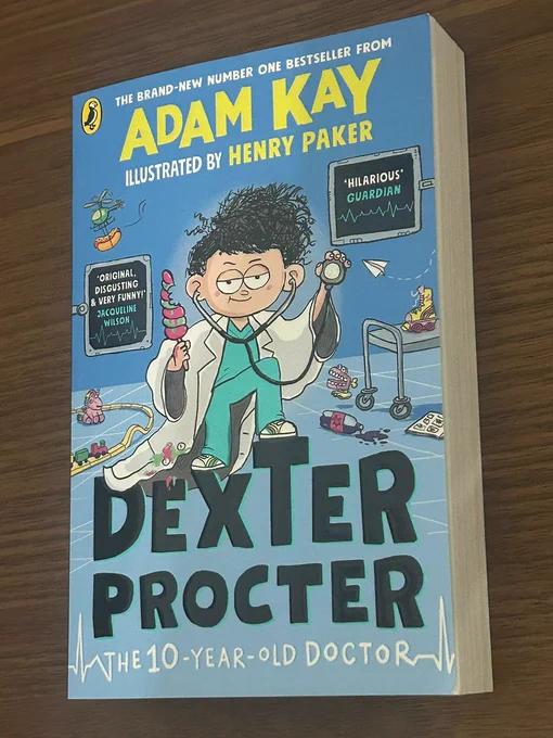 A blue book on a wooden table. DEXTER PROCTER THE 10-YEAR-OLD DOCTOR by Adam Kay, illustrated by Henry Paker. It features a picture of a child holding a stethoscope in a medical ward covered with toys and quotes "Hilarious" from The Guardian and "Original, Disgusting and Very Funny!" from Jacqueline Wilson.