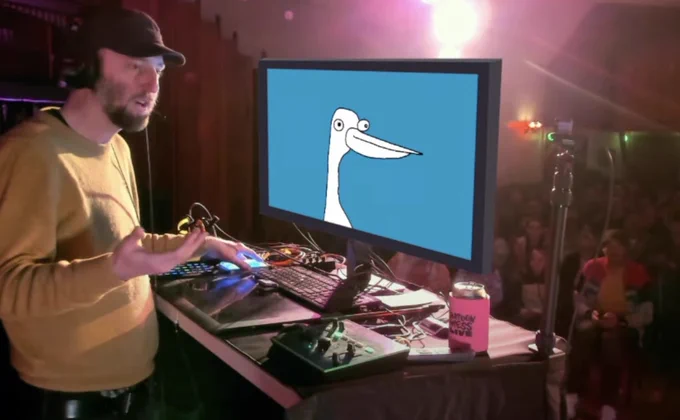 Tom DesLongchamp, wearing a cap and headphones, gestures next to a monitor displaying his animated character the Duck on a blue background, during a live show.  tomdeslongchamp.com
