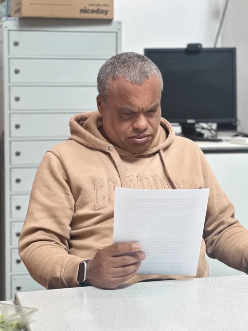 A middle-aged man with grey hair and wearing a brown hooded top is sitting at a table and reading a letter.