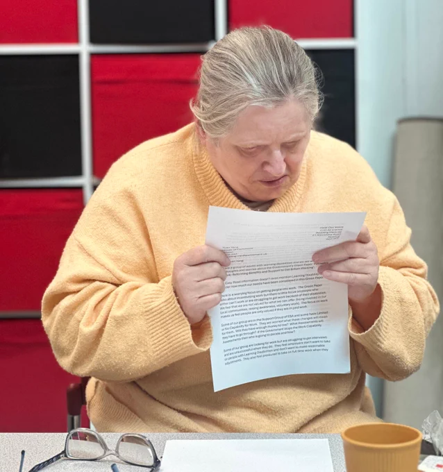 A middle-aged woman with blonde hair and wearing a yellow jumper is sitting at a table and reading a letter.