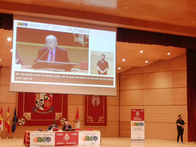 Imagen del ponente junto al moderador en la mesa del escenario durante la intervención. Ademas, del intérprete de lengua de signos.