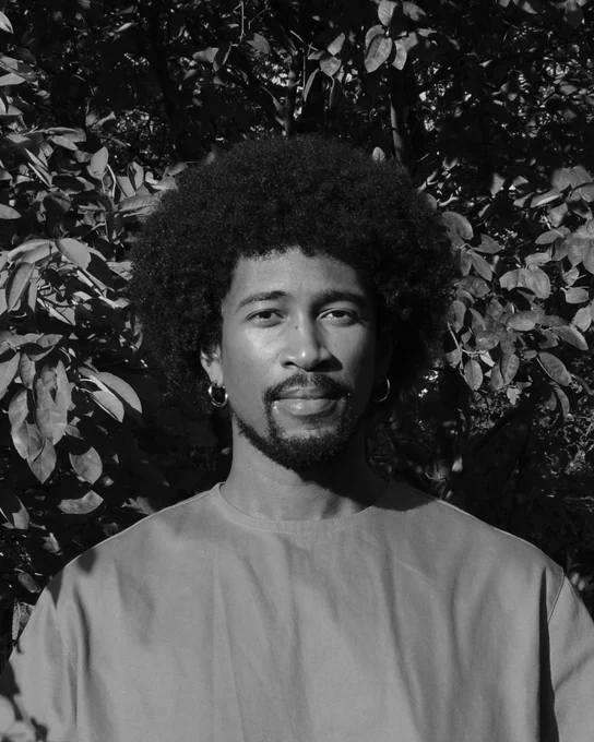 A black-and-white headshot of a Black person with an afro, goatee, and small hoop hearings. They are wearing a t-shirt, standing in front of a leafy background.
