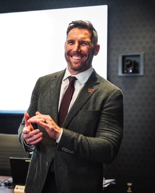 Photo of GM Adam Peters during the 2025 NFL Draft. Peters is smiling and clapping. He is wearing a gray suit with a burgundy tie and a white shirt underneath.