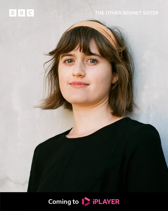 A woman in a black round neck top leans against a wall smiling. She wears a beige headband in her short brown hair.