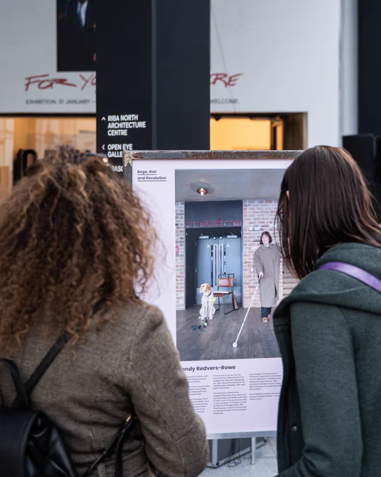 Two women are facing away from us, one with curly and one with straight hair. They are looking at a board in an exhibition with a photo of a woman walking with a white stick and a guide dog