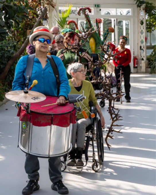 A procession of people move through Sefton Park Palm house. The person at the front is playing a drum. Behind them are people displaying various sculptures of disability aids pimped up with foliage and thorns