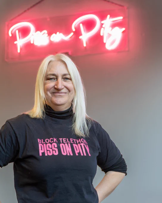 Zoe Partington stands in front of a Neon sign reading 'Piss on Pity' in Red
