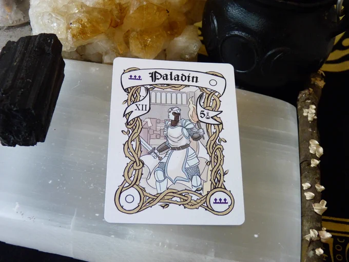 The Full Moon Paladin card has a full moon in 2 corners and a purple crown in the other two, with yellow border vines, and a Black man in white armor with a sword in one hand and a lit torch in the other. Behind him a table is on its side. In the banner there is the Roman Numeral XII on the left side and the Arabic number 5 on the right. The card is laying on a selenite slab, surrounded by a chunk of black tourmaline, a cluster of yellow citrine, a triple moon goddess cauldron, and a stick with lichen, all on a black and yellow cloth.