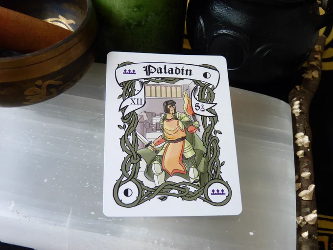 The Quarter Moon Paladin card has a quarter moon in 2 corners and a purple crown in the other two, with green border vines, and an Asian man in green and orange armor with a sword in one hand and a lit torch in the other. Behind him a table is on its side. In the banner there is the Roman Numeral XII on the left side and the Arabic number 5 on the right. The card is laying on a selenite slab, surrounded by a singing bowl, a green candle, a triple moon goddess cauldron, and a stick with lichen, all on a black and yellow cloth.