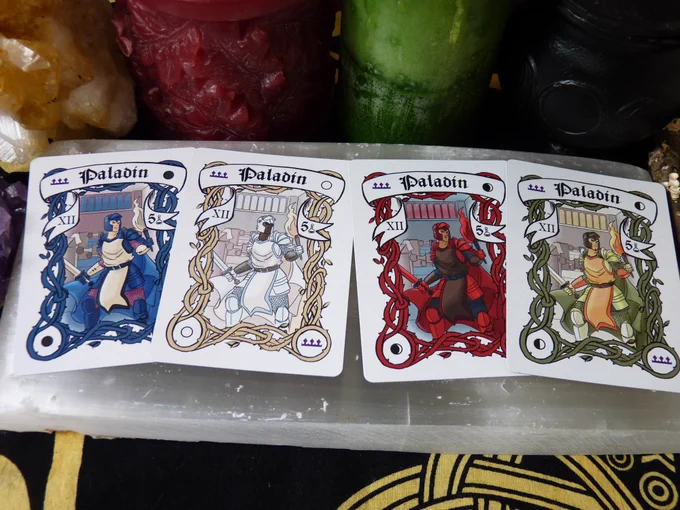 All 4 Paladin cards are laying on a selenite slab in order of New, Full, Crescent, and Quarter, surrounded by a cluster of purple amethyst, a cluster of yellow citrine, a red candle, a green candle, a triple moon goddess cauldron, and a stick with lichen, all on a black and yellow cloth.