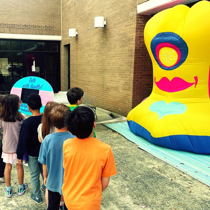 Line of kids wait to talk to a yellow alien inflatable. 