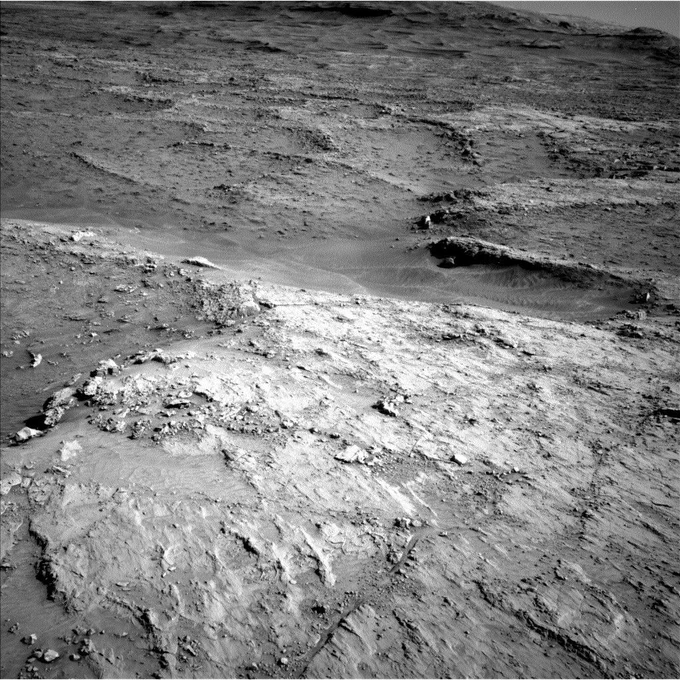 A ribbed Martian landscape is shown in a black-and-white image taken by the Curiosity rover. The long fractures stretch out toward the horizon, appearing jagged and cracked. 
