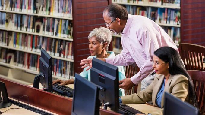 An image of two women using computers in a library whilst a man helps them. 