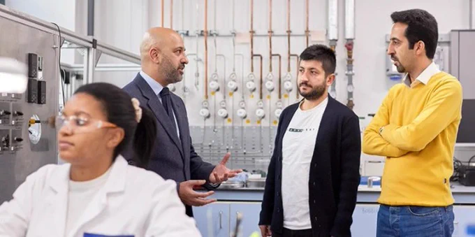 Three individuals listen to a fourth person, who is speaking and gesturing, in a laboratory setting filled with scientific equipment.