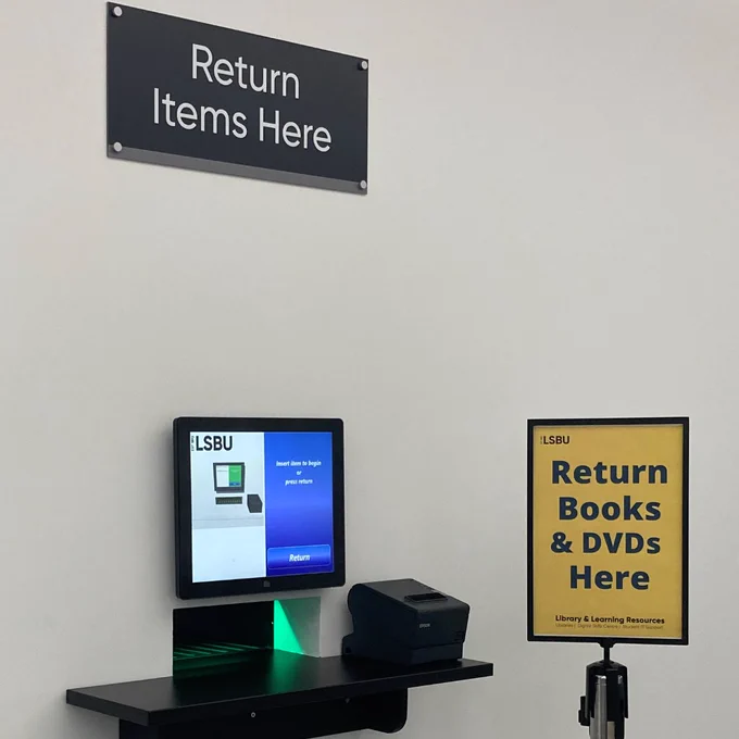 Photo of the book returns machine on level 03 of the Southwark Campus Library, LSBU Hub. Black sign above it that reads 'Return Items Here' and yellow poster in a stand in front of it that reads 'Return Books & DVDs Here'