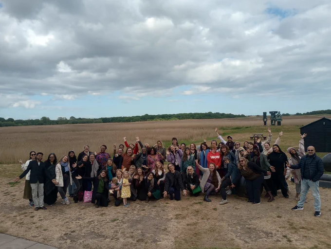 A group image of about 49 delegates on the Arts Health Research Intensive 2025. Behind the smiling group of delegates are black sculptures, reeds and a blue sky. 
