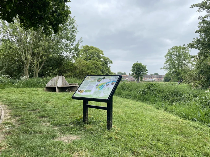 An open area of grass mown by one of the volunteers with tall vegetation retained at the edge. One of three interpretation boards funded by the 2022/23 Blue Green Corridor Project with the “Compass Bench” in the background. The bench was installed some 20 years ago, constructed by a Bourne Grammar School student with Oak timber extracted from Chambers Wood, Lincolnshire