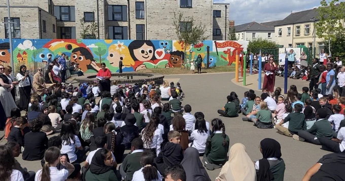 A large group of school pupils, in the playground, with a new mural on a wall in the background. Grŵp mawr o ddisgyblion ysgol, yn y maes chwarae, gyda murlun newydd ar wal yn y cefndir.