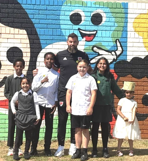 A group of school pupils stand, with an adult in front of a mural, on a wall. Mae grŵp o ddisgyblion ysgol yn sefyll, gydag oedolyn o flaen murlun, ar wal.