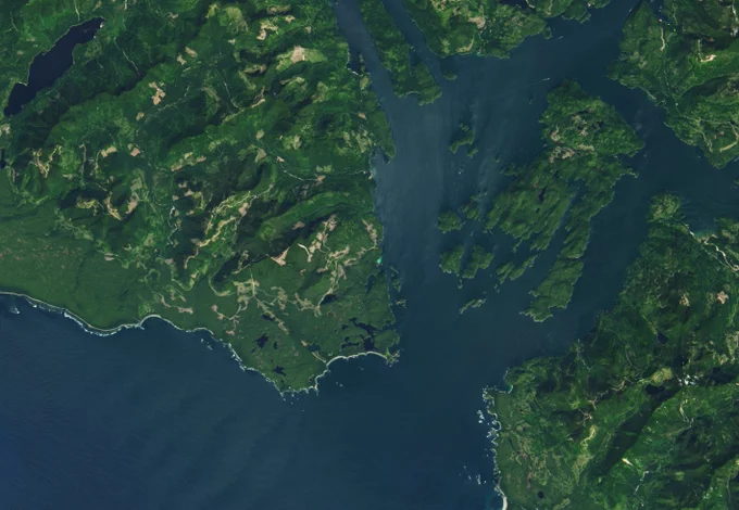 Satellite image showing a coastline with many islands and inlets. The land is lush green and mountainous, surrounded by dark blue waters. In one area, right next to the coastline, there is a bright turquoise glow in the water.